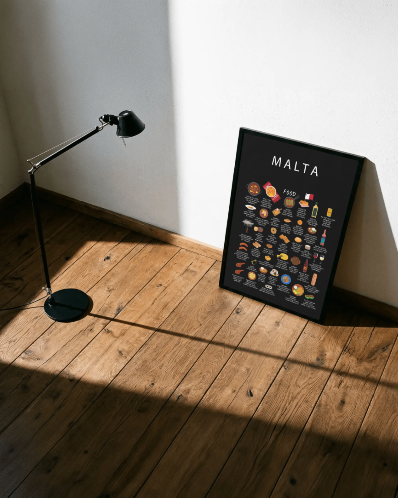 Framed black Malta food poster leaning against a sunlit wall on a wooden floor