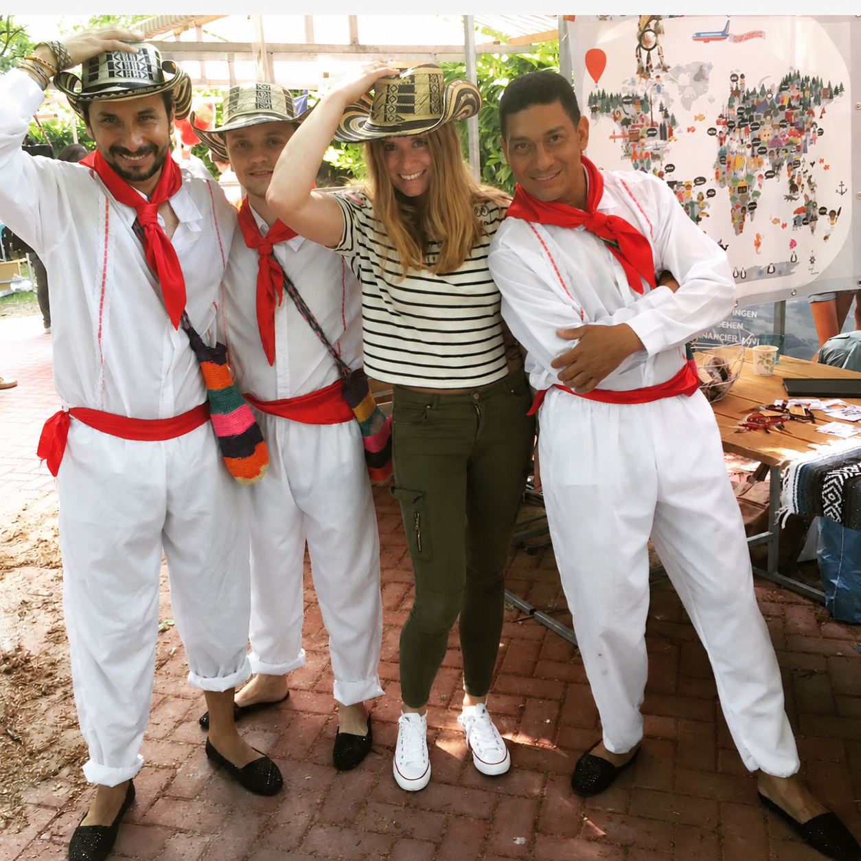 Anneke smiling beside three Colombian performers in white outfits, red sashes and woven sombreros vueltiaos in front of the hand-illustrated Maps By Annie world map at Wereldkinderdag in Wassenaar, 2015.