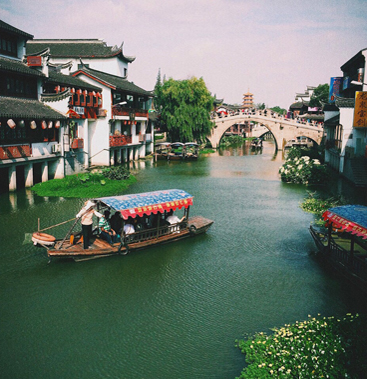 Traditional wooden boats with striped canopies gliding along a canal in the Zhujiajiao water town near Shanghai, lined with white-washed houses and a stone arch bridge.