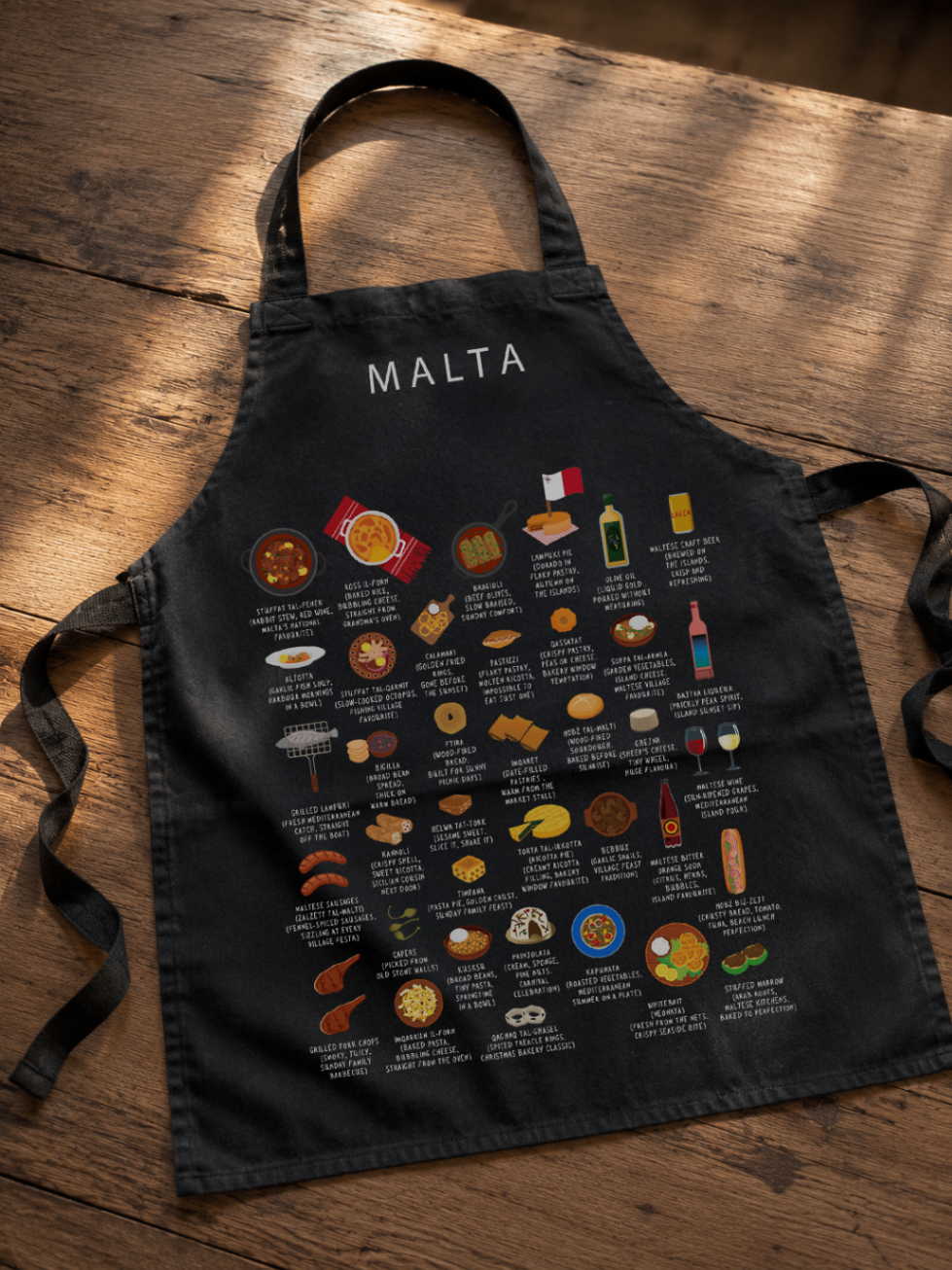 Black Malta food apron laid across a rich wooden floor in warm light