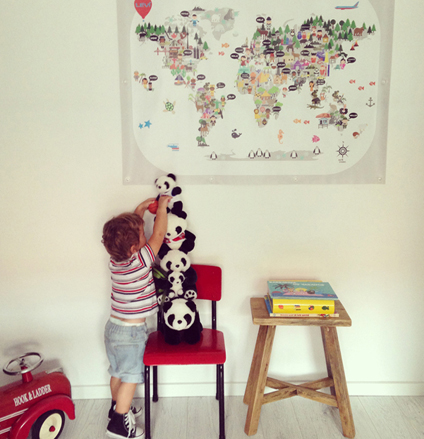 A young child stacking panda soft toys on a red chair beneath a large Maison Maps illustrated world map hanging on the wall of a bright nursery.