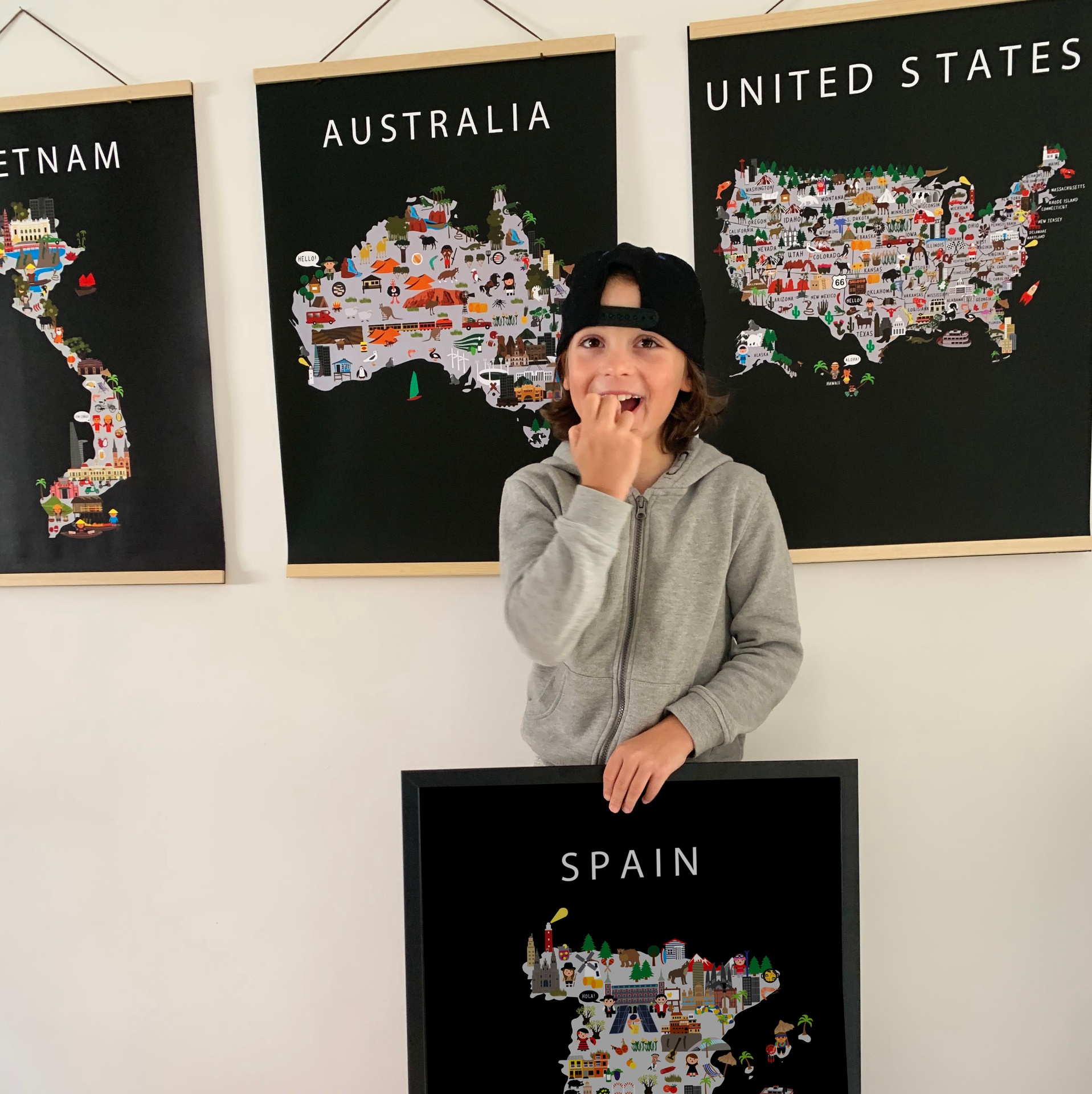 A boy standing in front of a gallery wall of framed Maison Maps posters for Vietnam, Australia and the United States while holding the Spain poster in front of him.