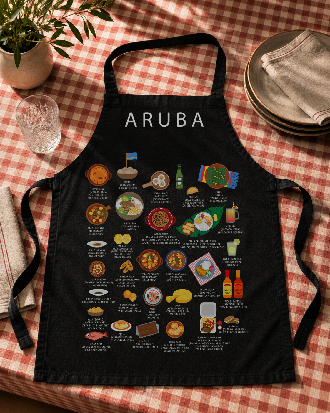 Black Aruba food apron flat-laid on a red gingham tablecloth with ceramic plates and a linen napkin