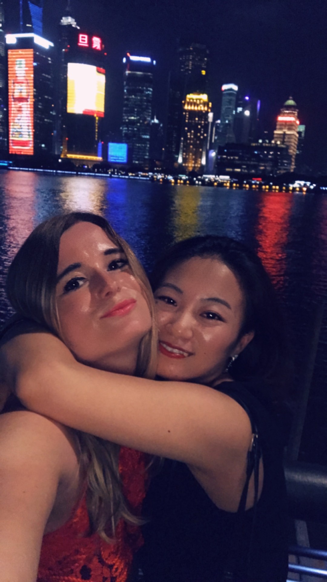 Anneke and her friend JiaJia embracing on the Shanghai Bund at night, with the illuminated Pudong skyline glowing across the Huangpu river behind them.