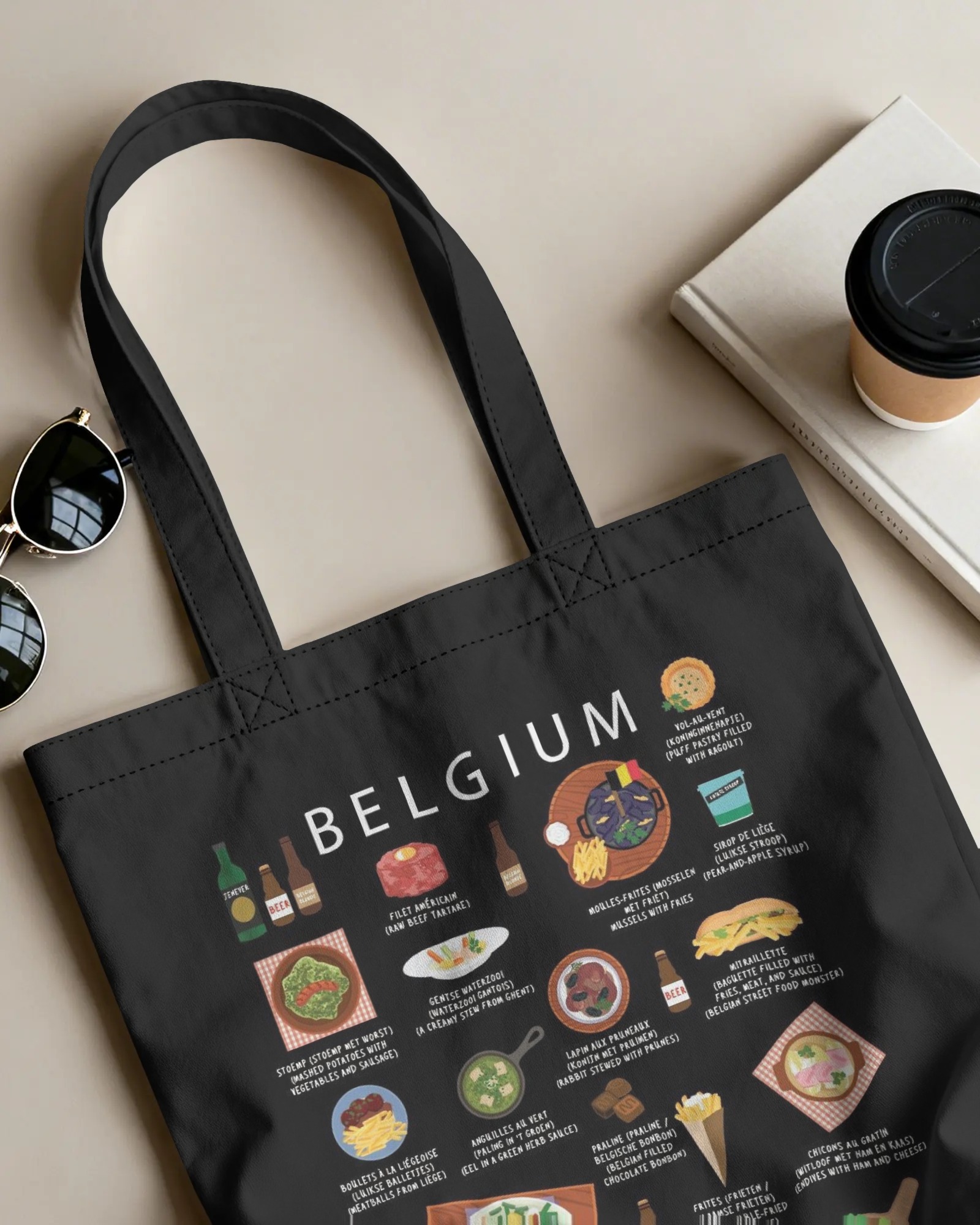 Black Belgium food tote bag laid flat beside sunglasses and a coffee cup on a neutral desk