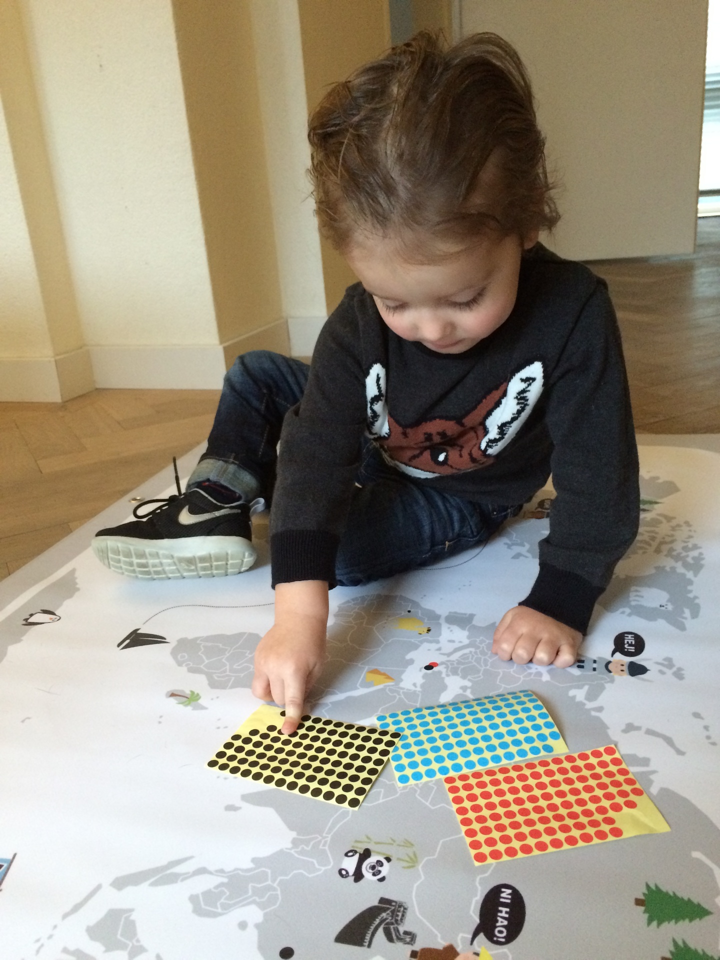 A young boy sitting on a wooden floor placing coloured dot stickers onto the European section of an early Maps By Annie illustrated world map.