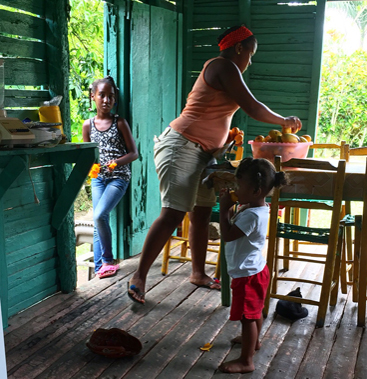 A family gathered around a wooden table on a bright green Caribbean porch, a scene that later inspired Maison Maps food illustrations.