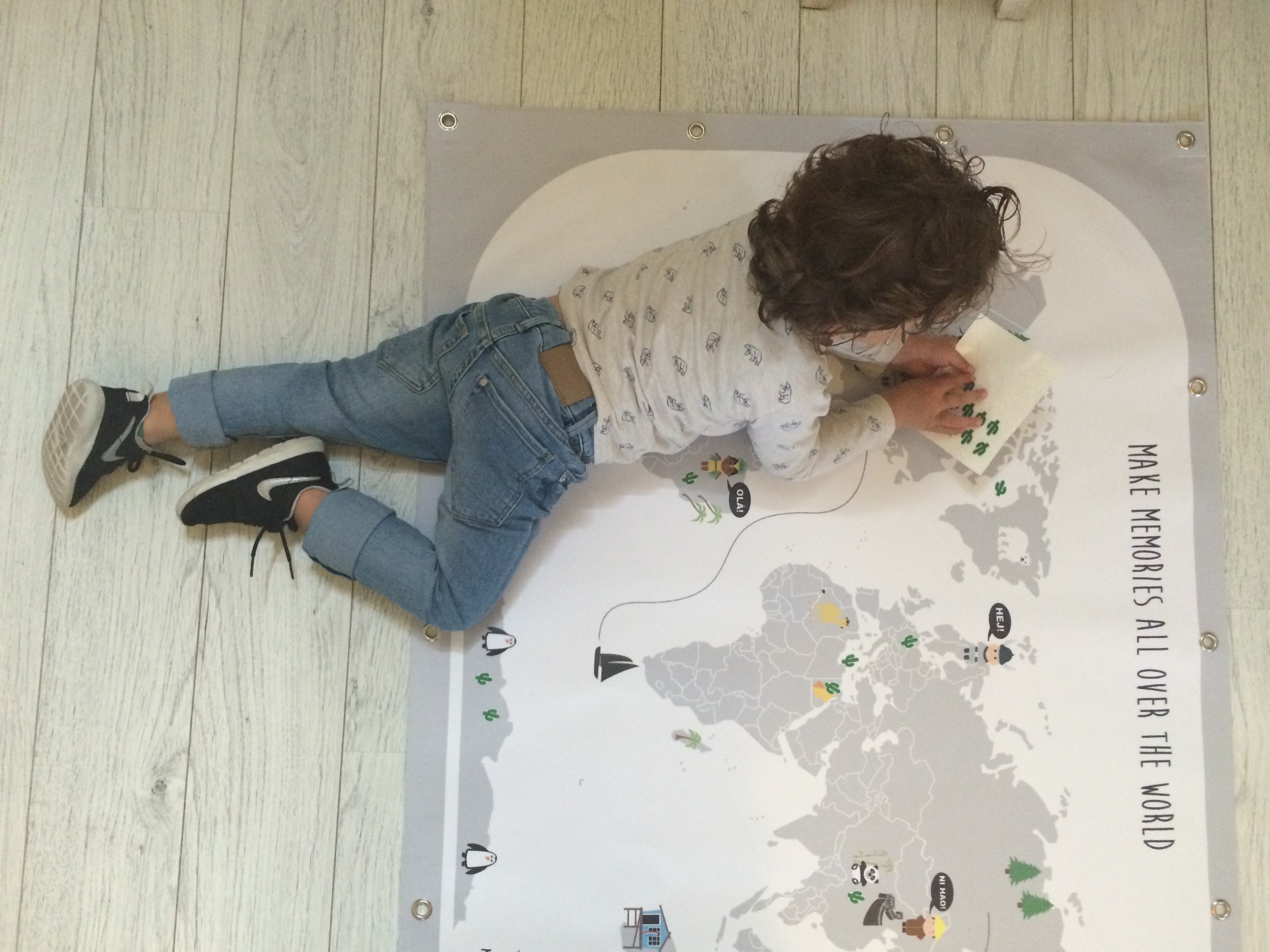 Overhead view of a young boy stretched across a large Maison Maps illustrated world map on a pale wooden floor, adding cactus stickers to the continents, with the words Make Memories All Over The World printed along the edge.