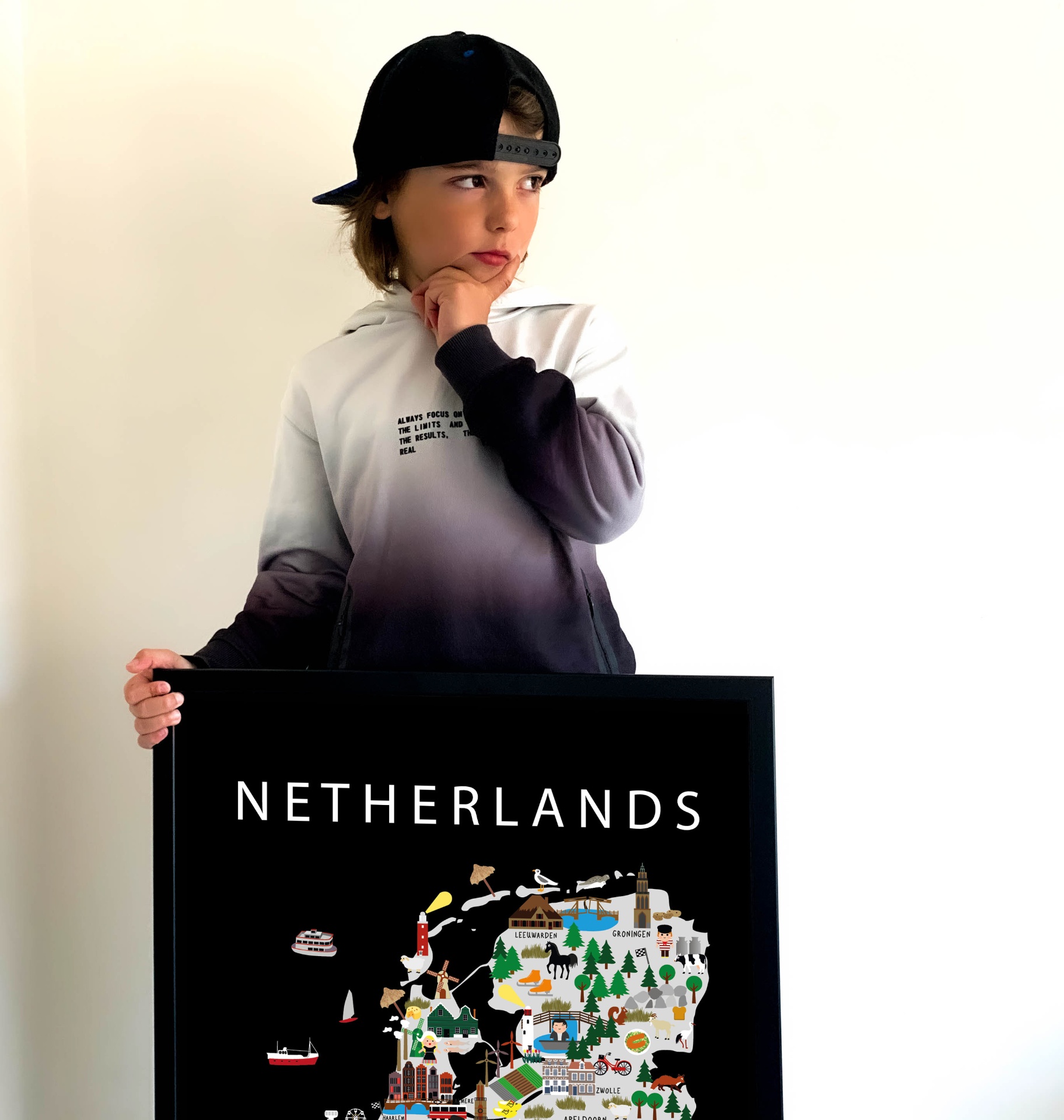 A boy in a black cap and gradient hoodie holding a framed Maison Maps Netherlands food and culture poster against a soft neutral wall.