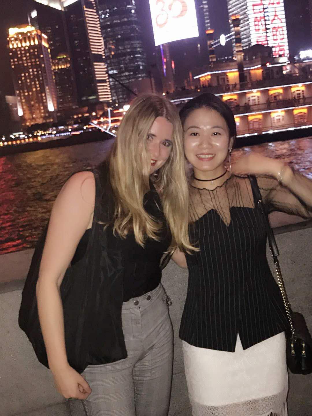 Anneke and JiaJia standing arm in arm on the Shanghai Bund in the evening, dressed up with the illuminated riverfront buildings behind them.