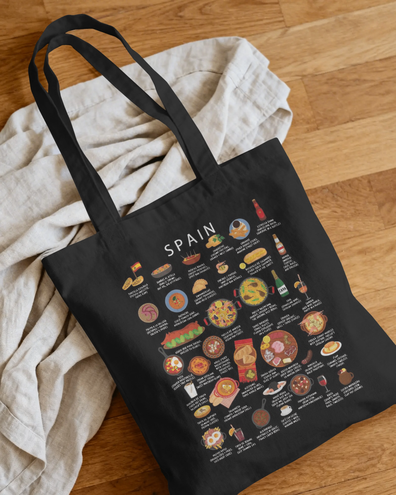 Maison Maps Spain illustrated black tote bag styled on linen and warm wooden flooring.