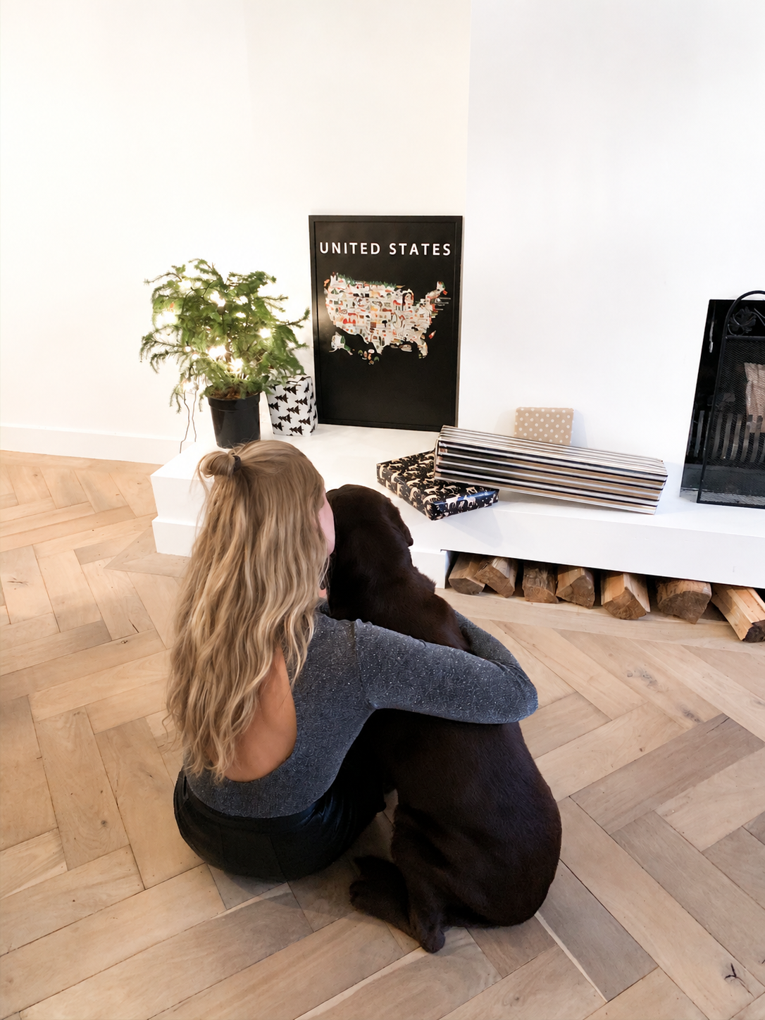 Anneke, founder of Maison Maps, sitting on the herringbone wooden floor with her dog Lewis, facing the framed Maison Maps United States poster in their home.