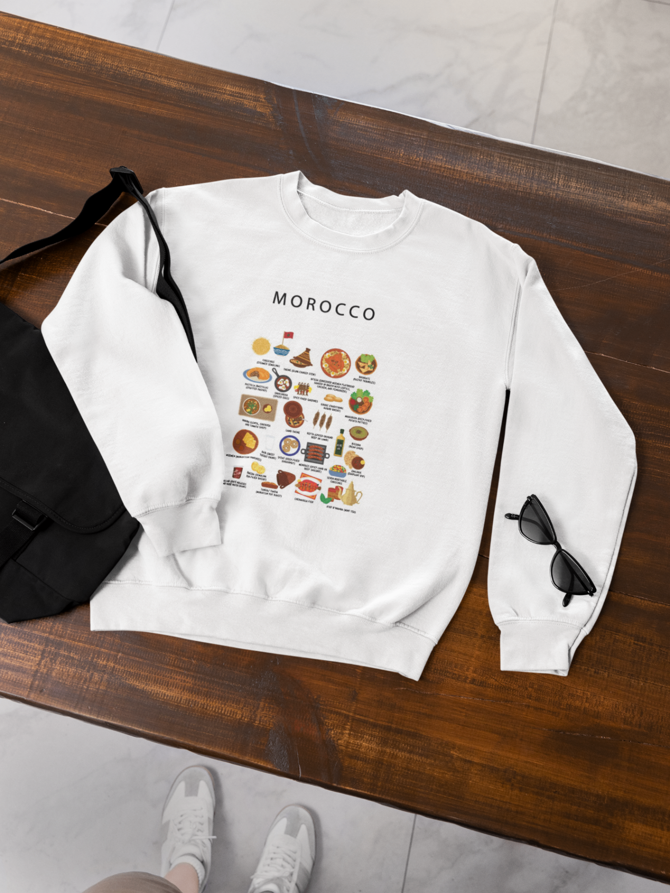 White Morocco food sweatshirt laid flat on a dark timber bench with a canvas bag and sunglasses