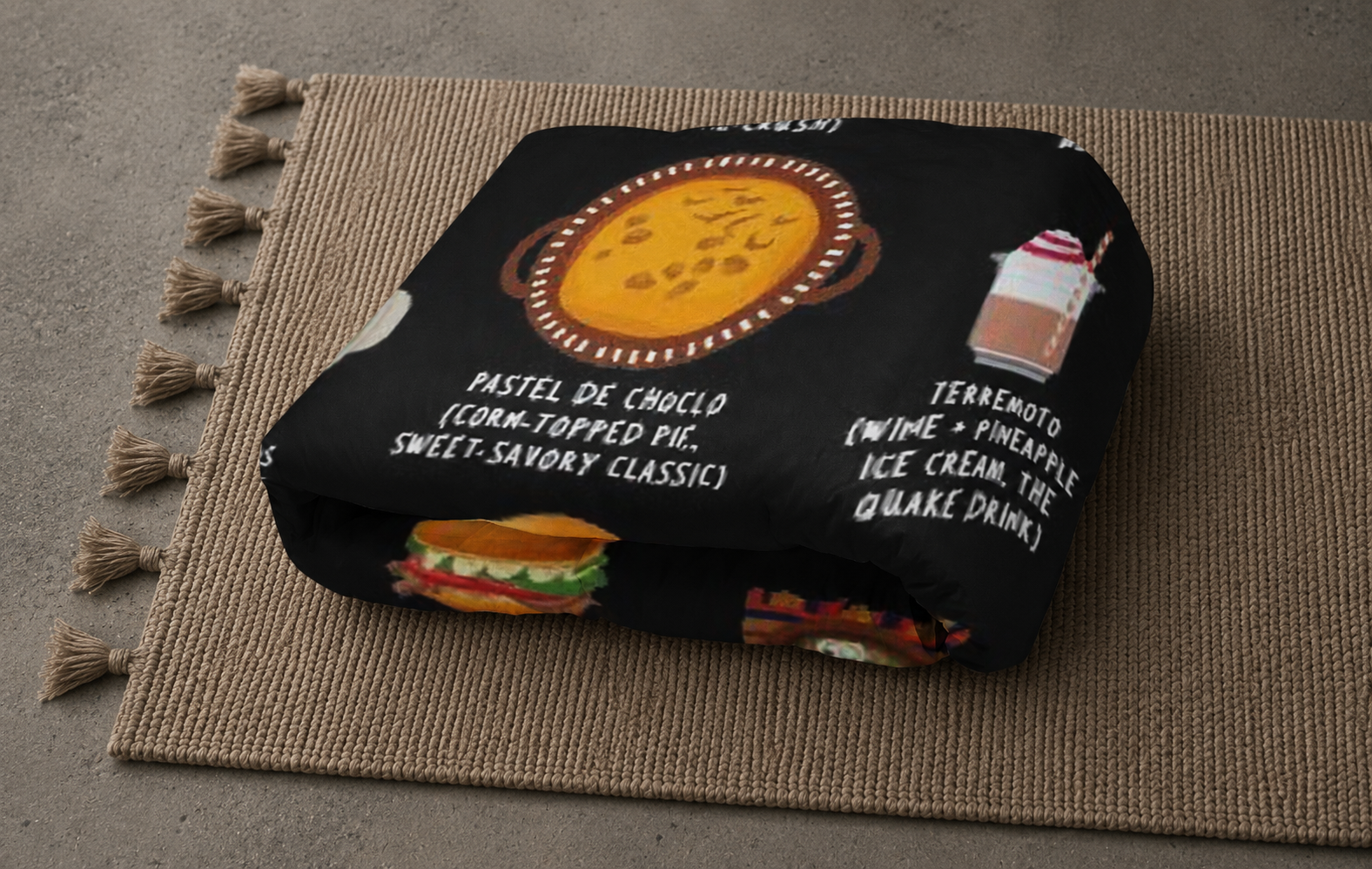 Folded black Chile food blanket on a woven jute mat, showing pastel de choclo and terremoto illustrations