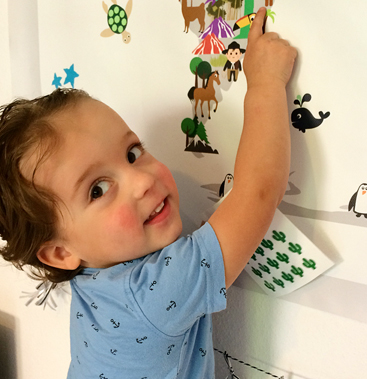 A smiling toddler in a blue anchor-print shirt pressing a sheet of cactus stickers onto a Maison Maps illustrated world map on the wall.
