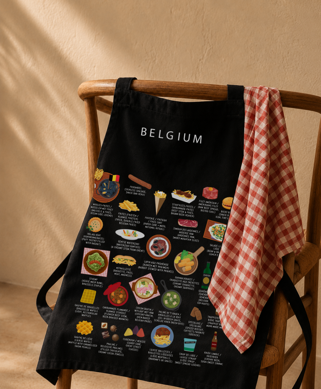 Black Belgium food apron hung on a wooden wishbone chair with a red gingham tea towel in warm afternoon light