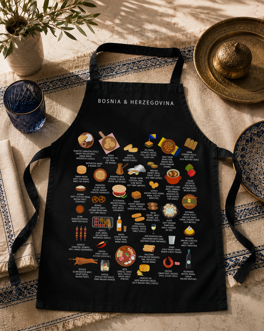 Black Bosnia & Herzegovina food apron laid on a woven mat beside a brass tray and blue glass in soft daylight