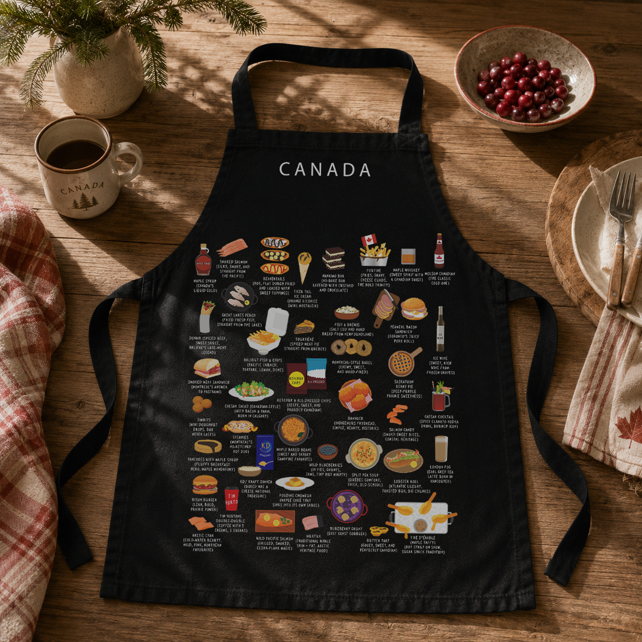 Black Canada food apron laid on a wooden table with a Canada mug, cranberries and cutlery