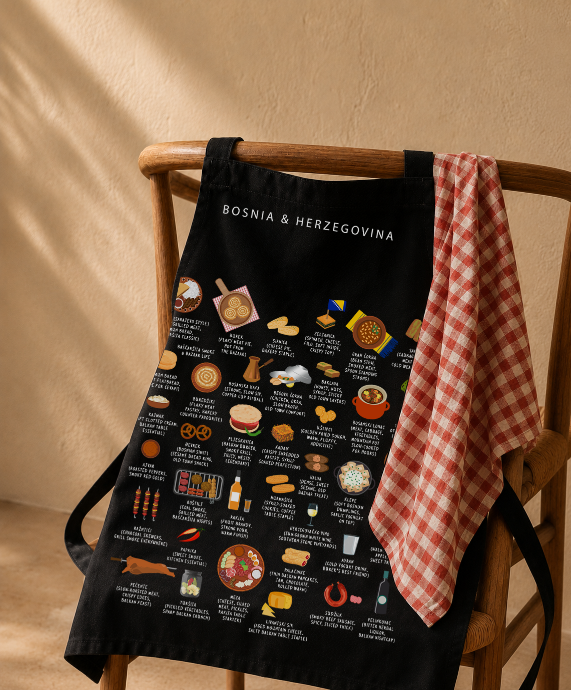 Black Bosnia & Herzegovina food apron hung on a wooden wishbone chair with a red gingham tea towel in warm afternoon light
