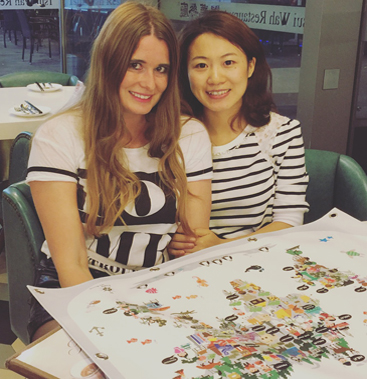 Anneke and her friend JiaJia sitting together behind a large hand-illustrated Maps By Annie world map at a restaurant in Shanghai, Anneke with long blonde hair and JiaJia in a black-and-white striped top.