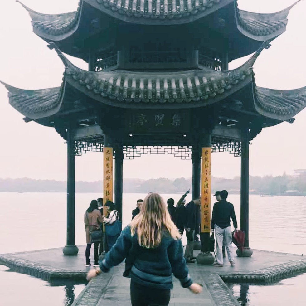 Anneke walking towards a traditional Chinese lakeside pavilion with sweeping tiled roofs, other visitors gathered under the pagoda on a misty day.