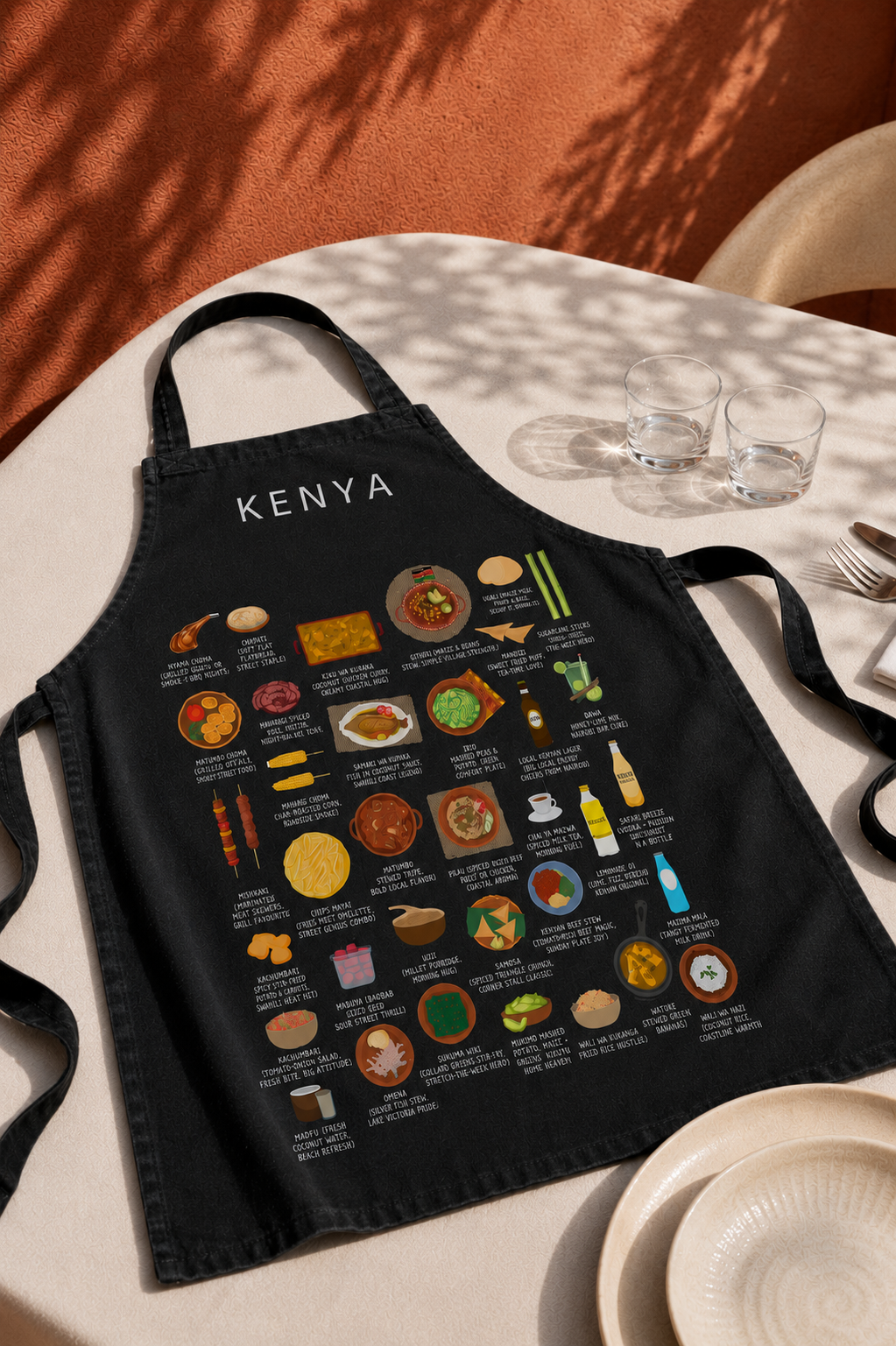 Black Kenya food apron on a sunlit outdoor table with terracotta wall, palm shadow and water glasses