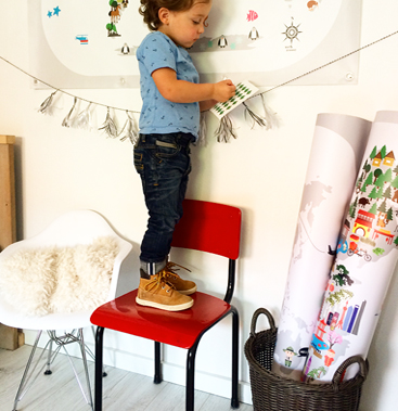 A small boy in a blue anchor-print shirt standing on a red chair to place illustrated stickers on a Maison Maps world map, with rolled poster tubes in a woven basket beside him.