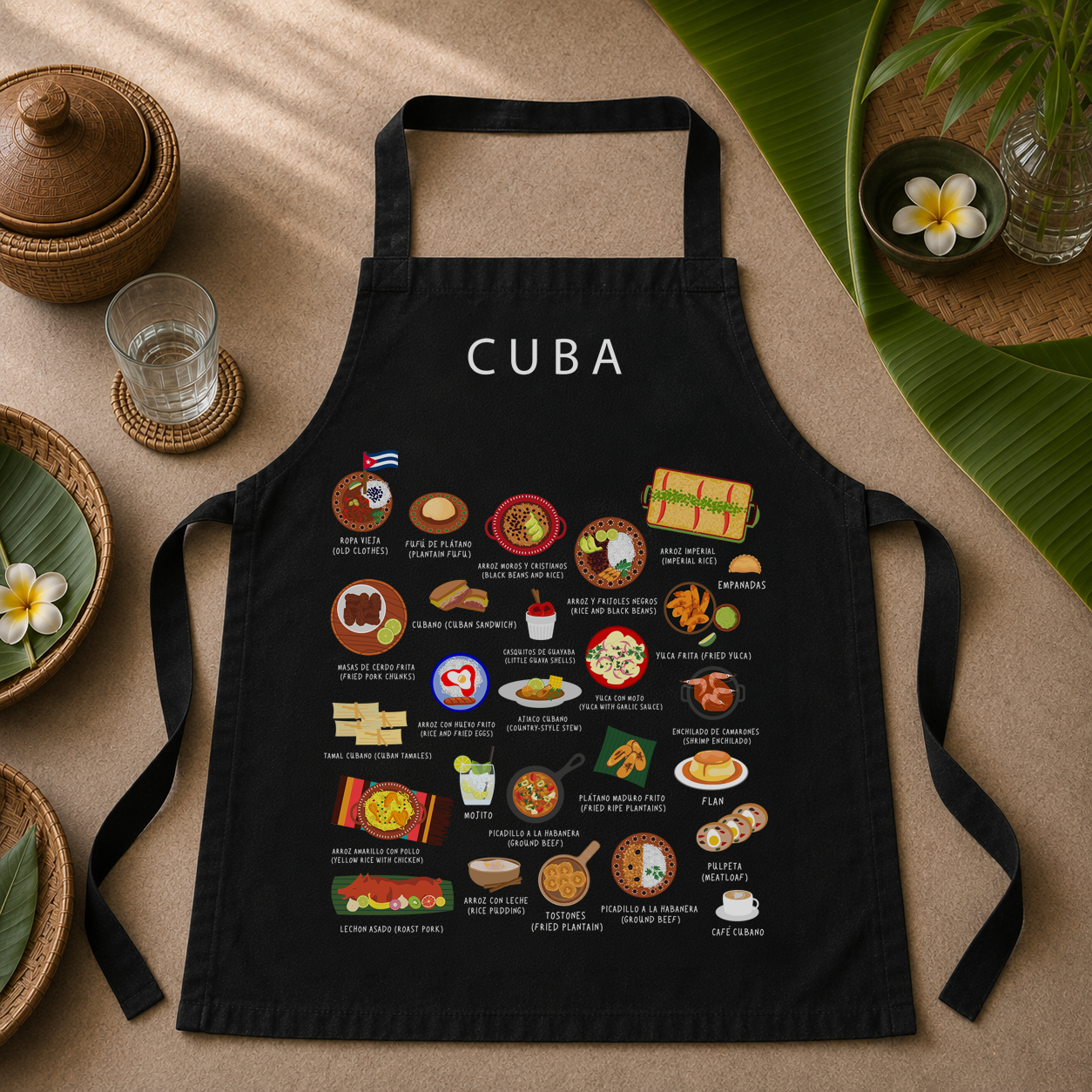 Black Cuba food apron styled flat on a warm sand-toned surface with woven baskets and palm leaves
