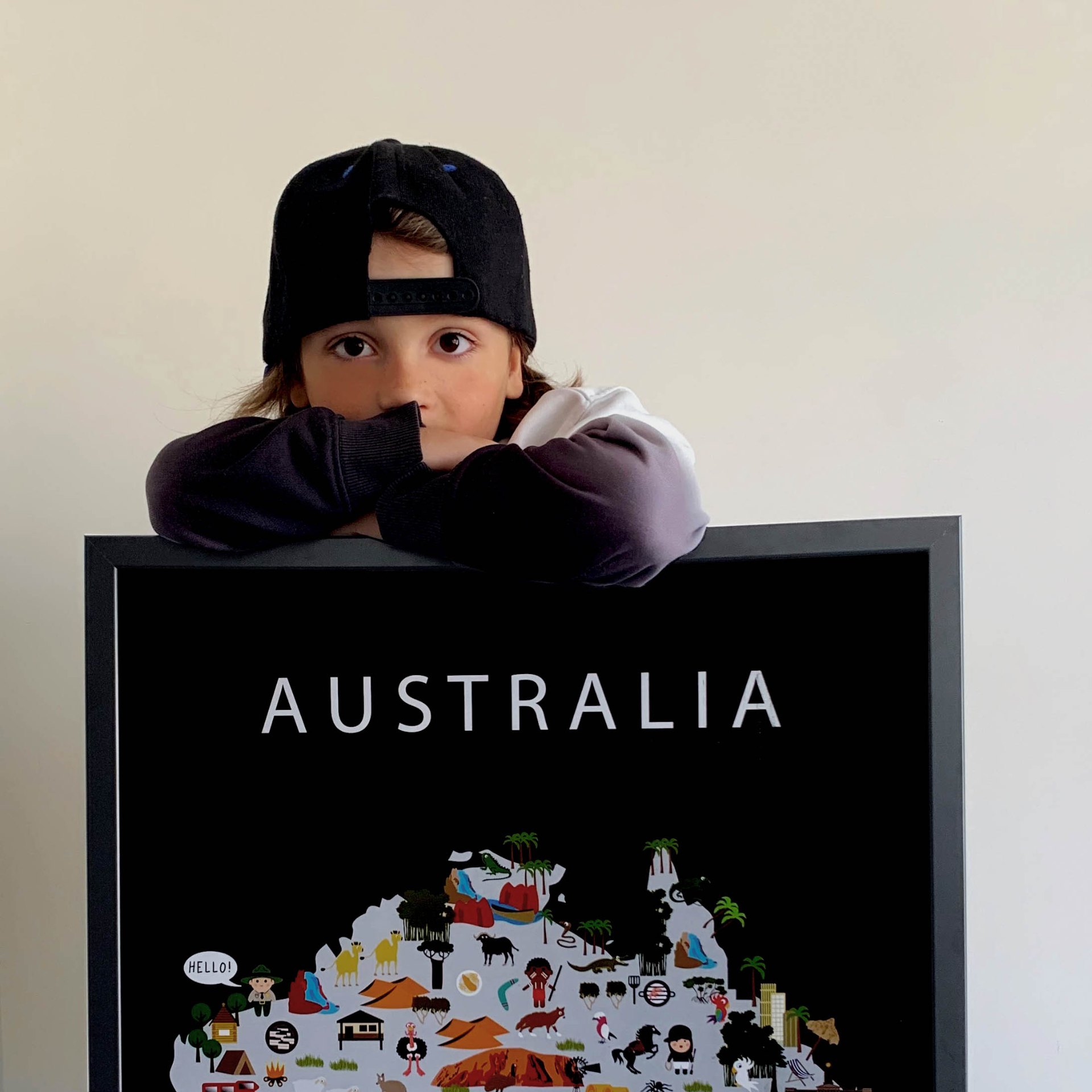 A boy in a black cap resting his arms on top of a large framed Maison Maps Australia food and culture poster in a bright, softly lit room.
